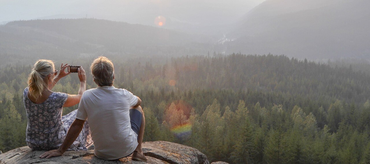 A couple seated on a cliff taking a photo of a mountain landscape