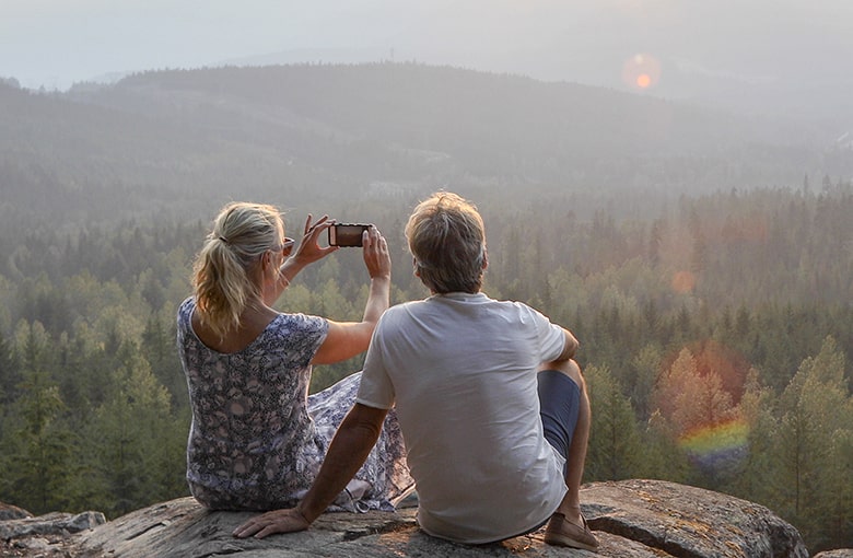 A couple seated on a cliff taking a photo of a mountain landscape