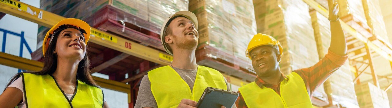Three young workers wearing safety vests and hard hats stand together in a warehouse