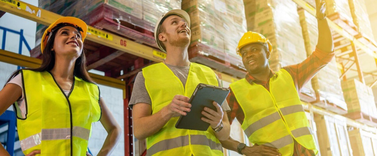 Three young workers wearing safety vests and hard hats stand together in a warehouse