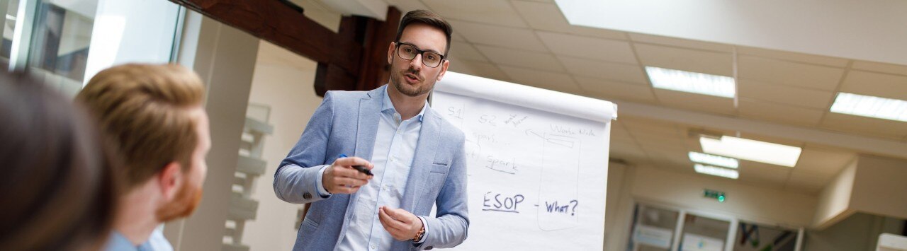 Man in suit in front of easel with large white paper, presenting in front of colleagues