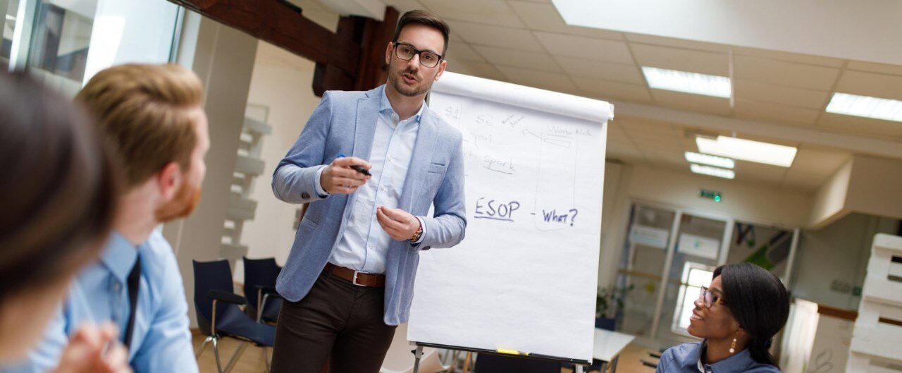 Man in suit in front of easel with large white paper, presenting in front of colleagues
