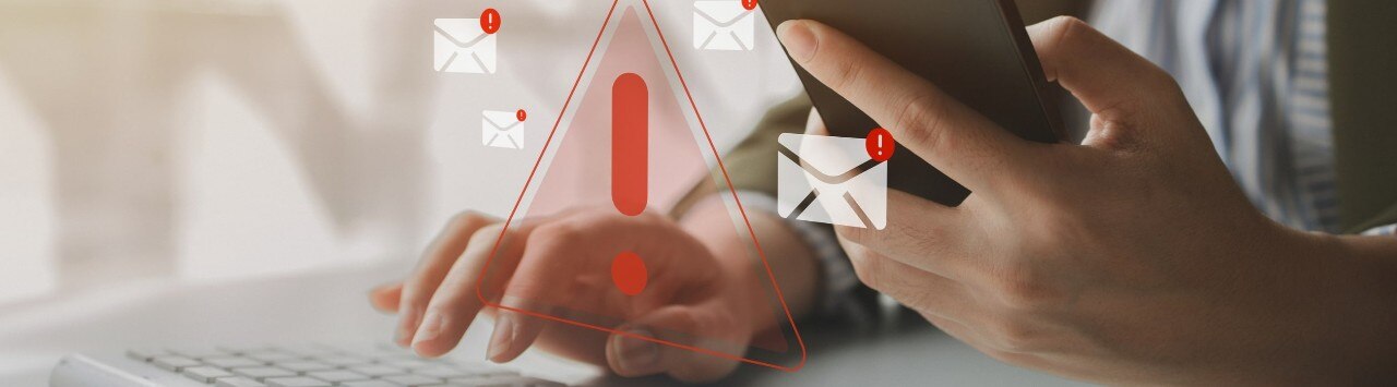 Close up of hands working with a laptop and smartphone, with superimposed graphics of email icons and red warning exclamation points