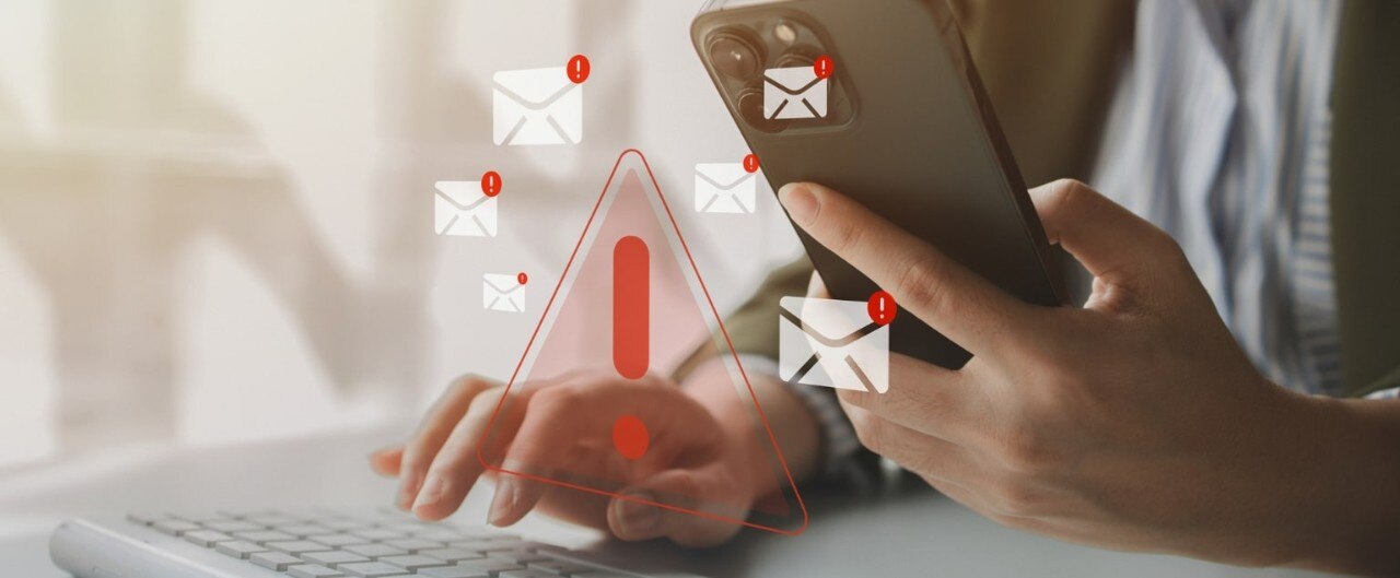 Close up of hands working with a laptop and smartphone, with superimposed graphics of email icons and red warning exclamation points