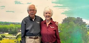 An elderly man and elderly woman stand with their arms around each other, smiling in front of a mural of their town. 