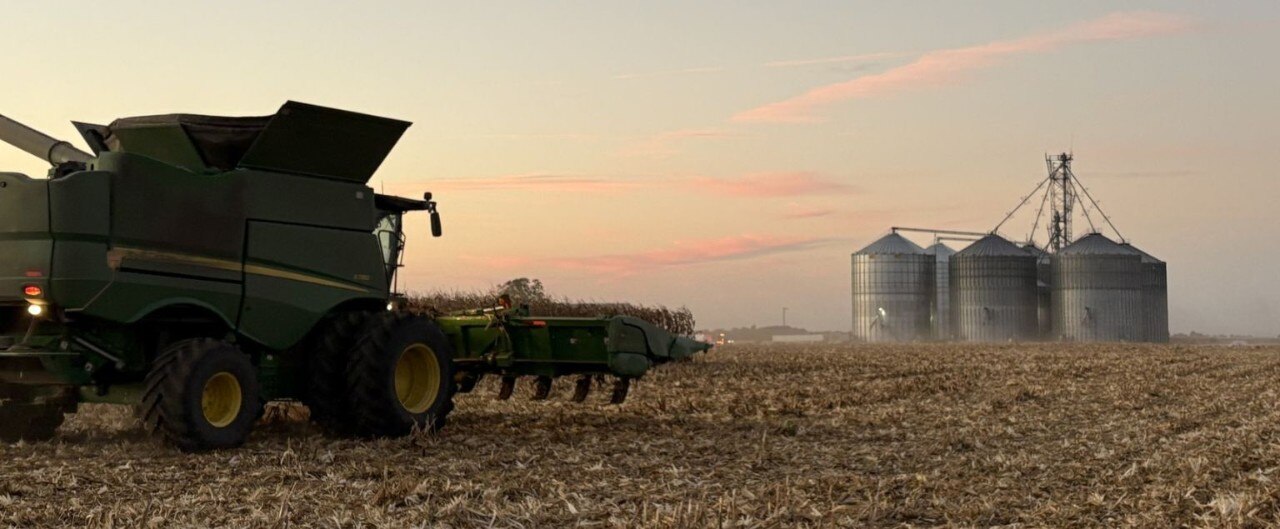 Wide shot of corn field at dusk with combine in foreground and grain elevators in background.