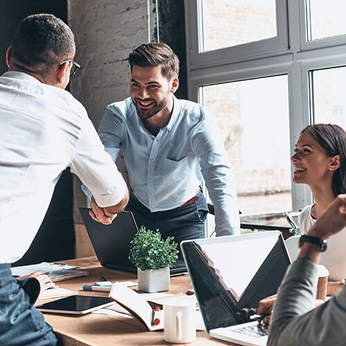 Two businesspeople shaking hands at a conference table as additional colleagues look on smiling