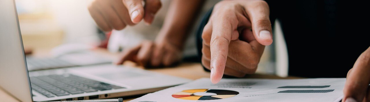 Two hands point at printed charts and a laptop, indicating a business discussion