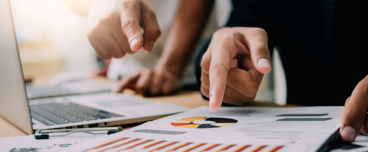 Two hands point at printed charts and a laptop, indicating a business discussion