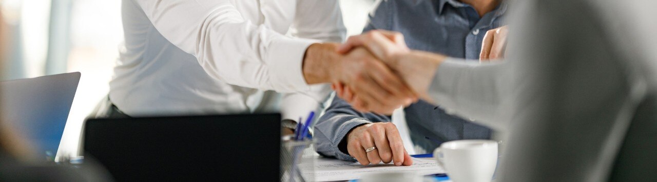Middle-aged business men shaking hands in a brightly lit office setting.