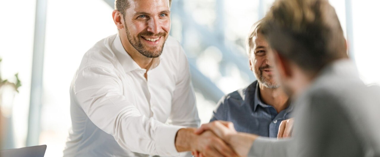 Middle-aged business men shaking hands in a brightly lit office setting.