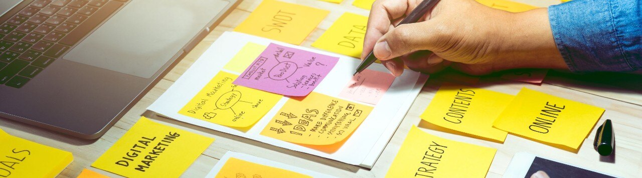 A laptop surrounded by colorful sticky notes across a desk, and hands writing more notes about marketing.