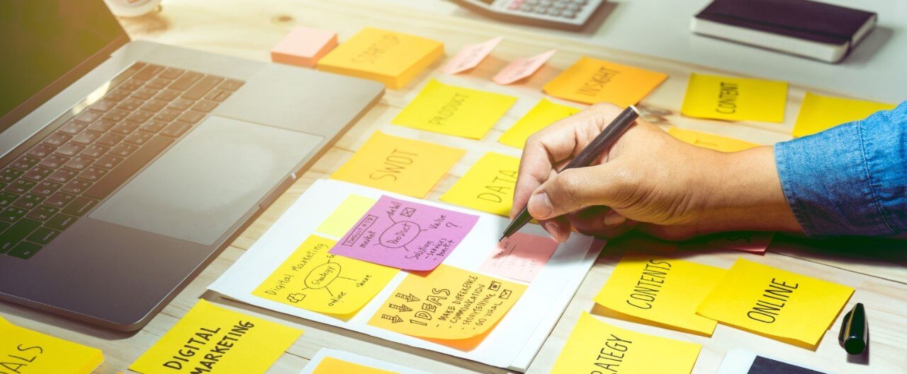 A laptop surrounded by colorful sticky notes across a desk, and hands writing more notes about marketing.