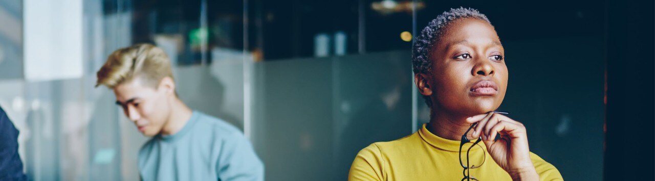 Businesswoman with close-cropped gray hair holds glasses under her chin and gazes into distance with concern