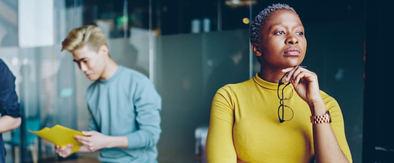 Businesswoman with close-cropped gray hair holds glasses under her chin and gazes into distance with concern