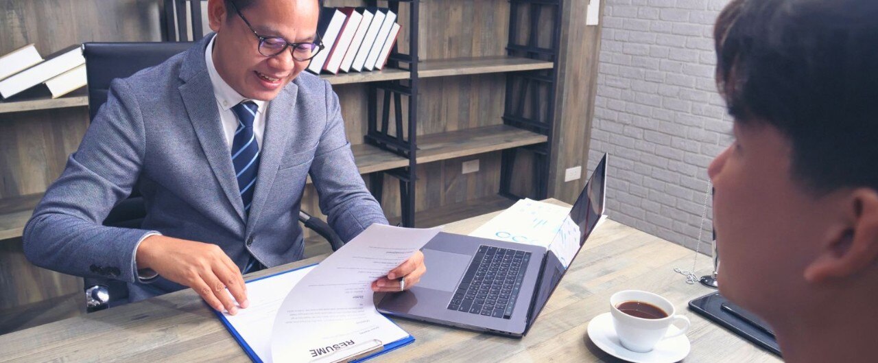 Man wearing glasses and a gray suit sits at a desk and reviews a resume while a younger man looks at him from across the desk. 