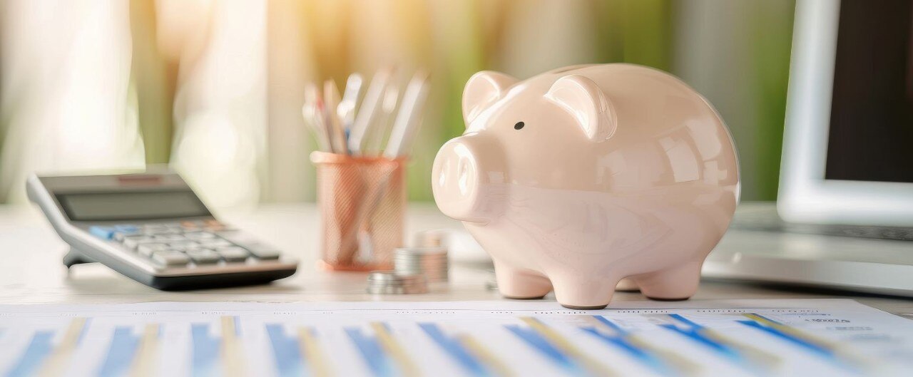 Desk with financial paperwork, calculator, pencil jar, stacks of coins and pale pink piggy bank.