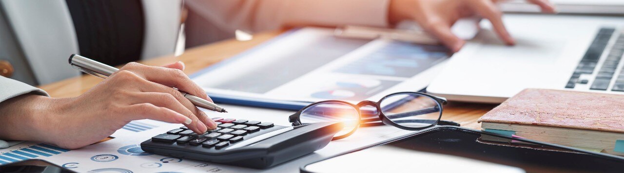 Detail view of desk covered with papers, charts, glasses, and a person’s hands working with a calculator and laptop.