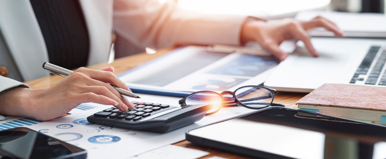 Detail view of desk covered with papers, charts, glasses, and a person’s hands working with a calculator and laptop.
