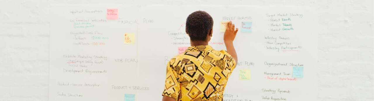 An African American person stands in front of a large whiteboard, organizing colorful notes and business strategy ideas.
