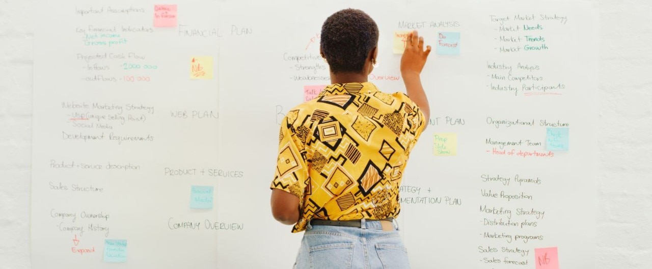 An African American person stands in front of a large whiteboard, organizing colorful notes and business strategy ideas.