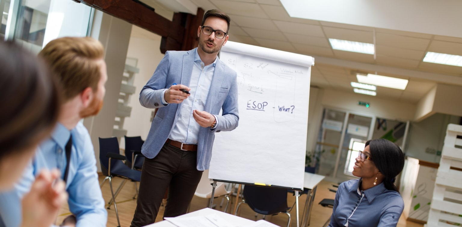 Man in suit in front of easel with large white paper, presenting in front of colleagues