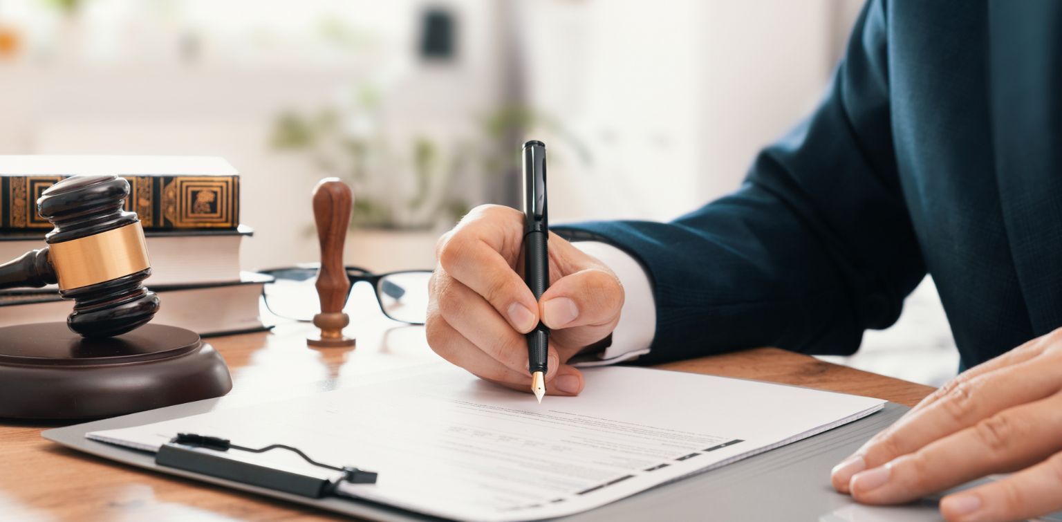A close-up of masculine hands signing an official document on a desk. A gavel, books, and glasses are on the desk in the background.