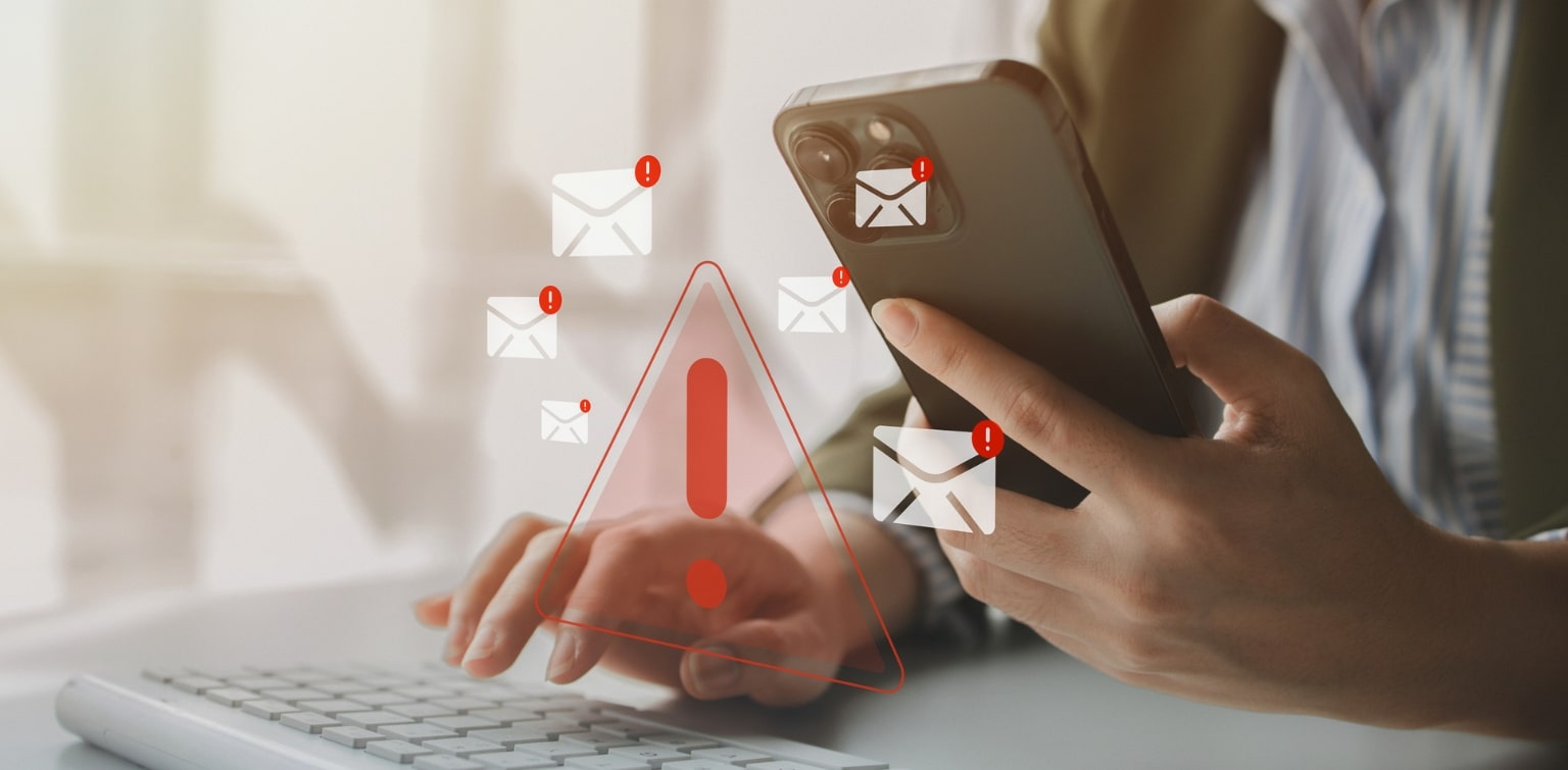 Close up of hands working with a laptop and smartphone, with superimposed graphics of email icons and red warning exclamation points