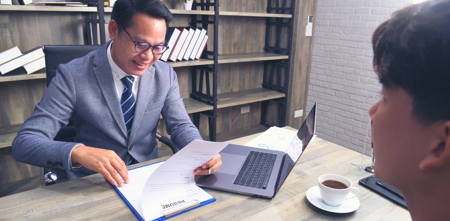 Man wearing glasses and a gray suit sits at a desk and reviews a resume while a younger man looks at him from across the desk. 