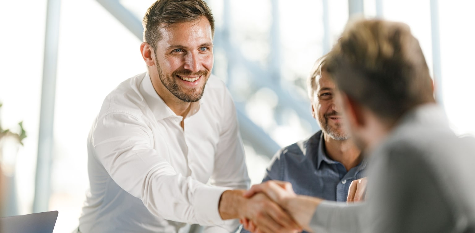 Middle-aged business men shaking hands in a brightly lit office setting.