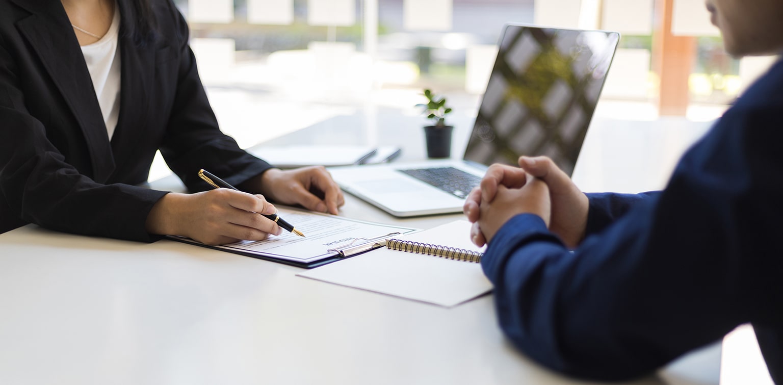 A man and a woman collaborate at a table with a laptop between them, discussing something on the screen.