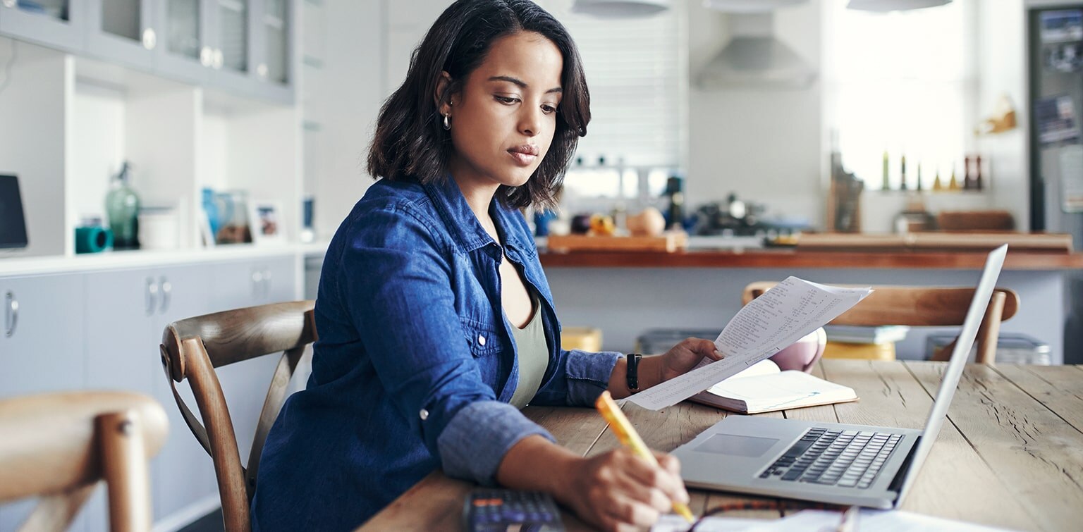 Shot of a young woman using a laptop and  going through paperwork while working from home