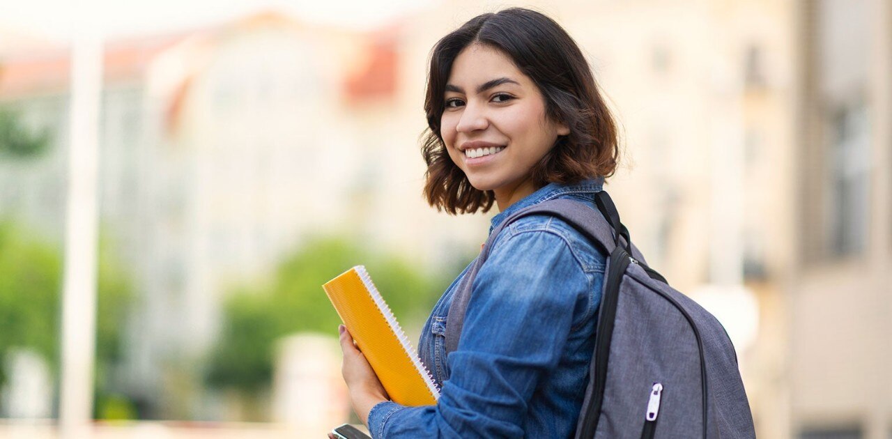 Smiling Hispanic female college student holding notebook, smartphone, and backpack