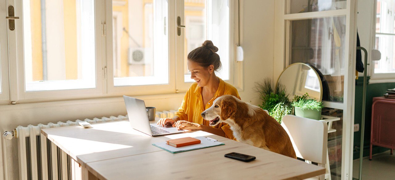 Young Caucasian woman smiling and working on laptop while her dog looks on