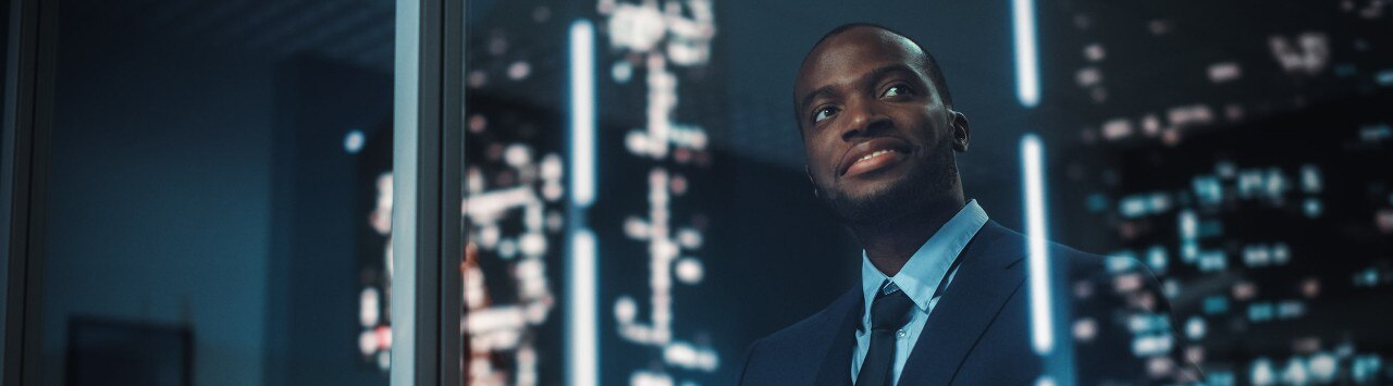 Happy, young African-American businessman looking out window at city at night