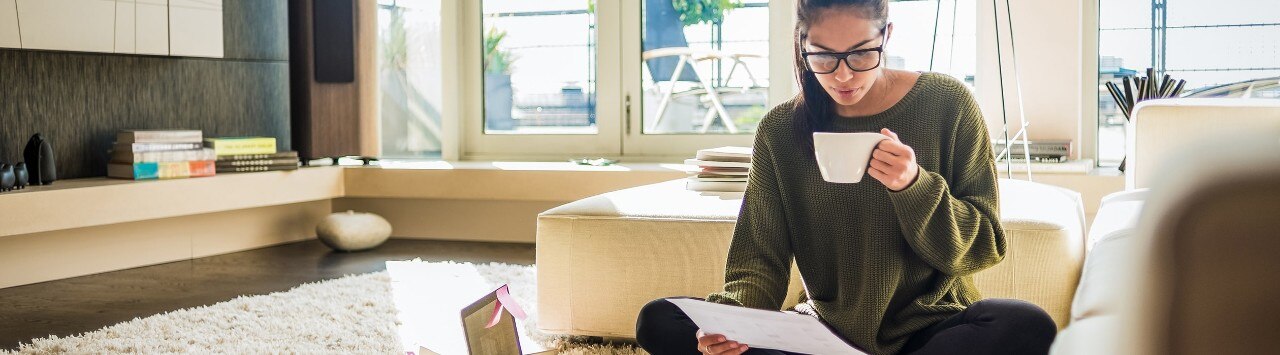 Young college grad sitting on floor at home reviewing bills and drinking coffee