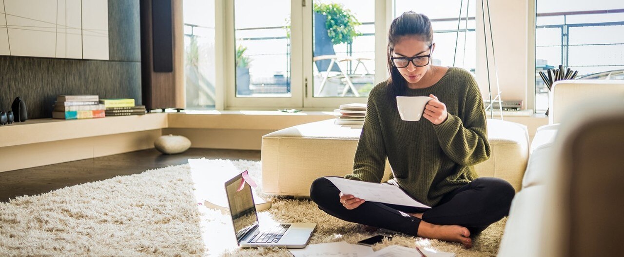 Young college grad sitting on floor at home reviewing bills and drinking coffee