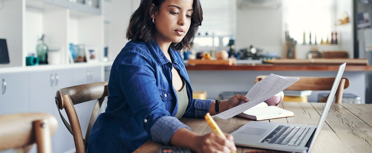 Shot of a young woman using a laptop and  going through paperwork while working from home