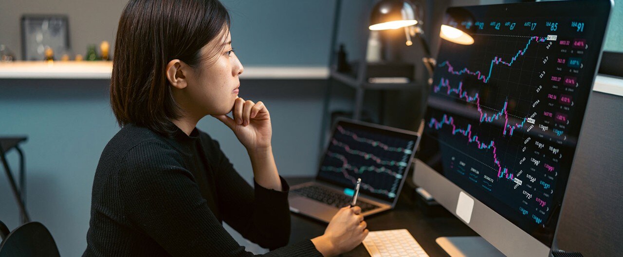 Asian-American female analyst reviewing stock market data on multiple monitors