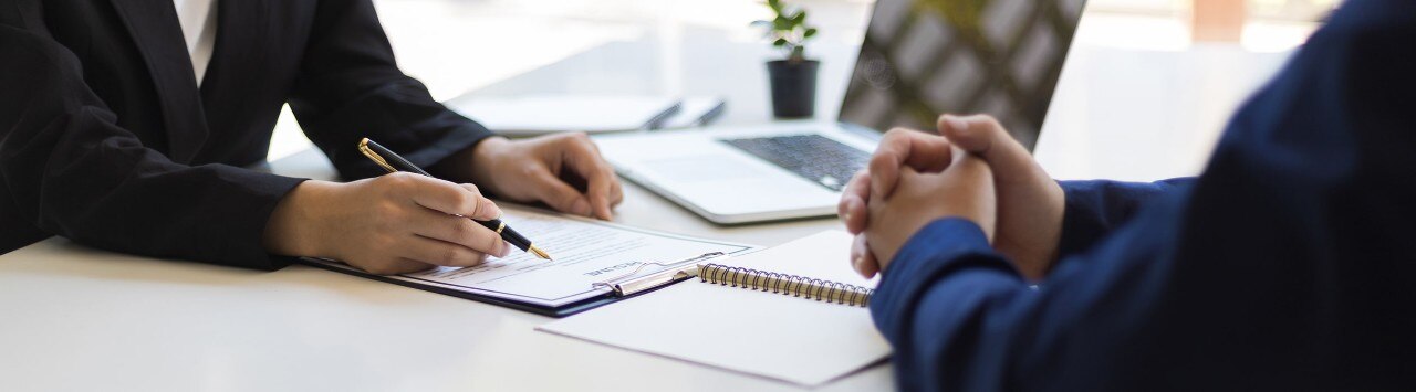 A man and a woman collaborate at a table with a laptop between them, discussing something on the screen.