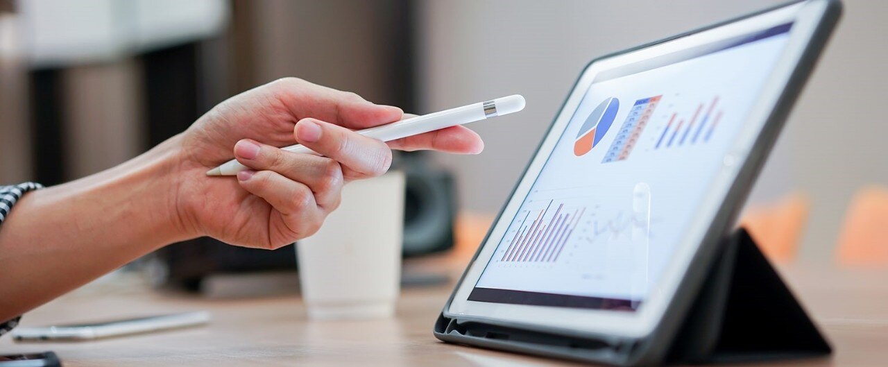 A hand pointing a stylus at a tablet screen displaying graphs and charts